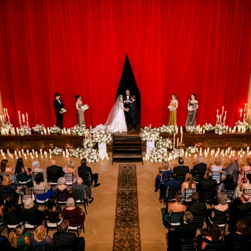 A wedding ceremony held in front of a large red curtain, with the bride and groom on stage surrounded by flowers and candlelight.