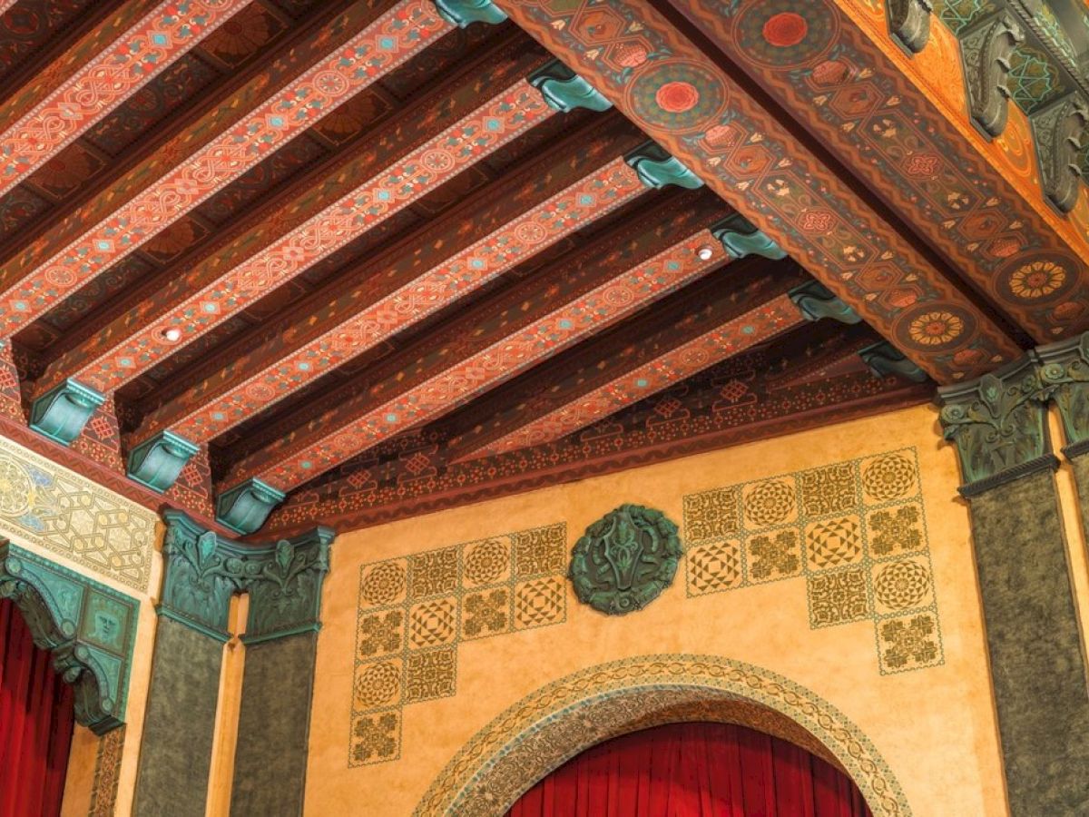 The image shows an ornate ceiling and wall with detailed patterns and arches, featuring rich colors and decorative elements.