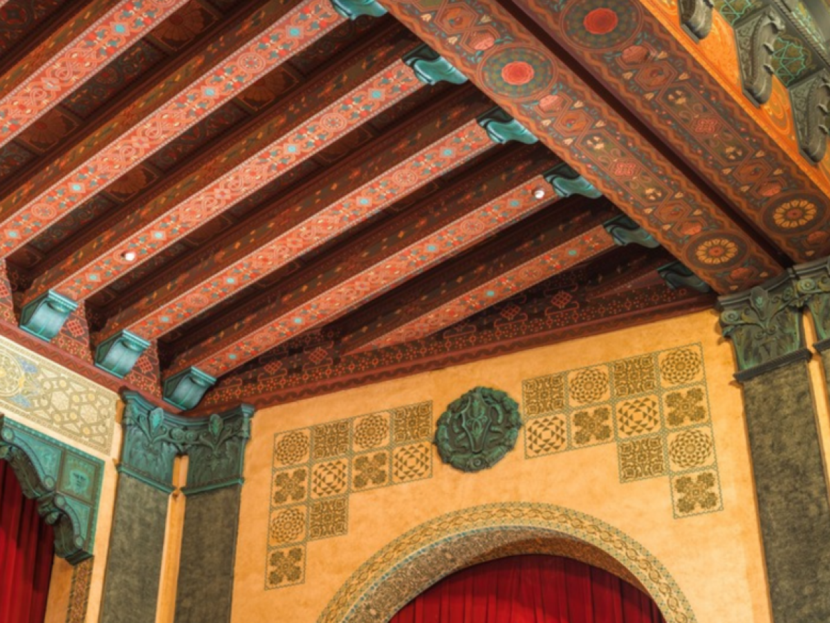 The image shows an ornate theater interior with intricate ceiling designs, arches, and red curtains.