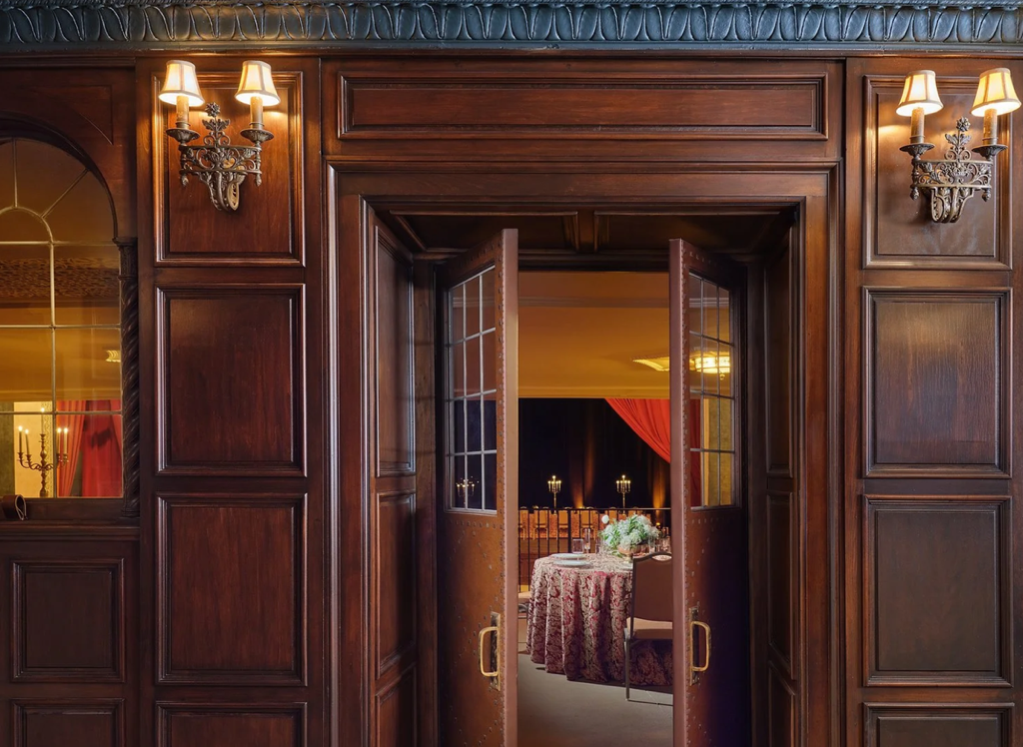 A wooden doorway with open doors leads to a dimly lit room, featuring a round table with a floral centerpiece and draped red curtains.
