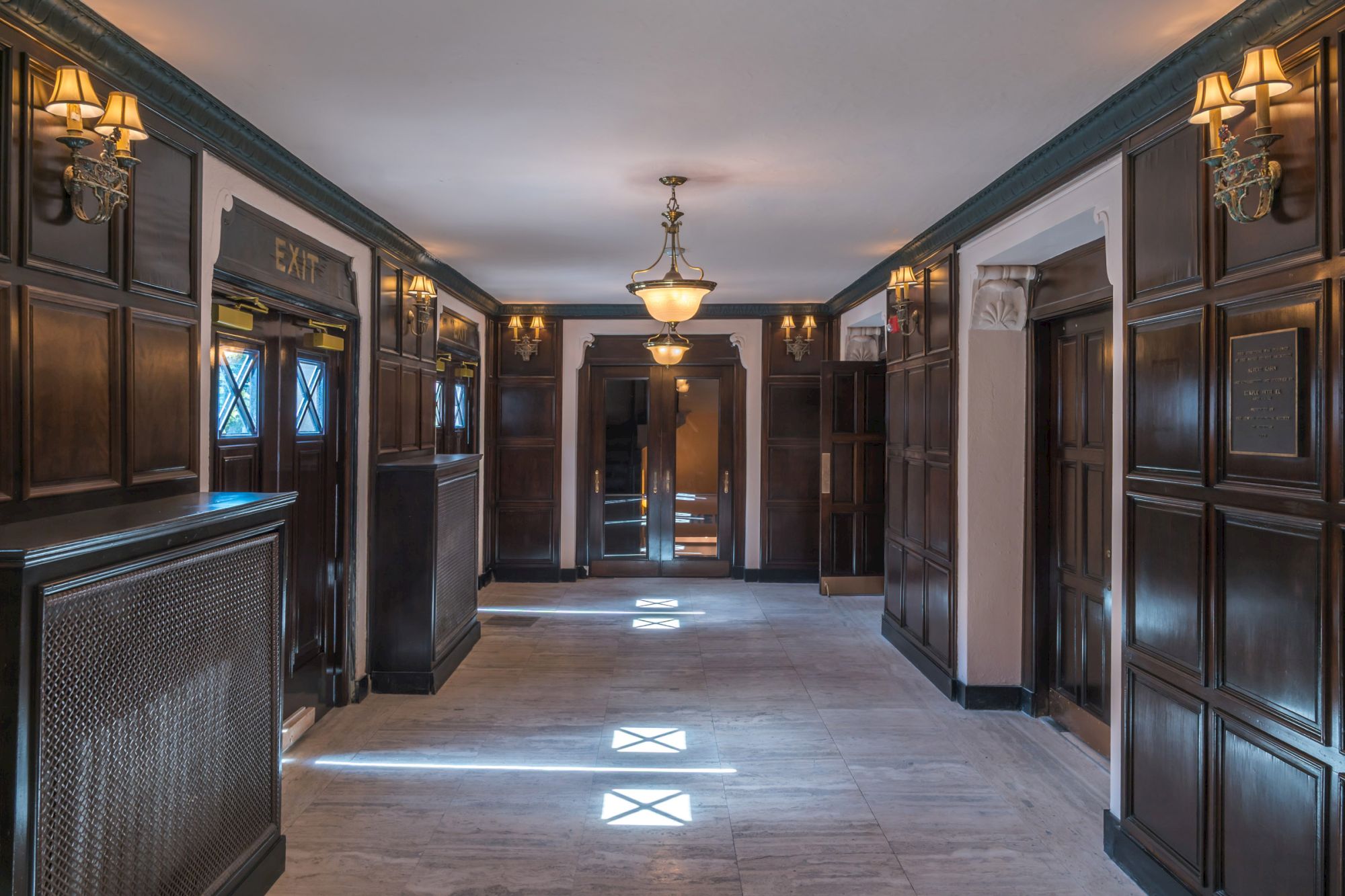 A hotel hallway with wooden paneling, chandeliers, and three elevators at one end, creating a classic and elegant ambiance.