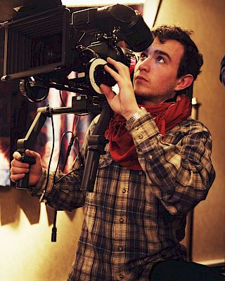 A person is operating a professional camera, focusing intently. They're wearing a plaid shirt and a red scarf in a dimly lit setting.