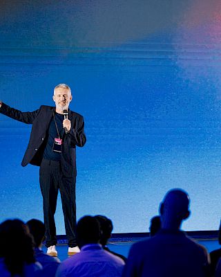 A person is speaking on a stage with a microphone, addressing an audience against a blue background.