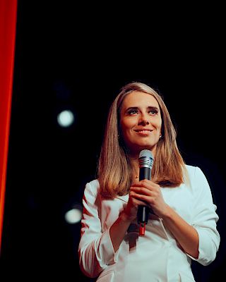 A person holding a microphone stands on stage with a red curtain and dark background, appearing to be in a speaking or performance event.