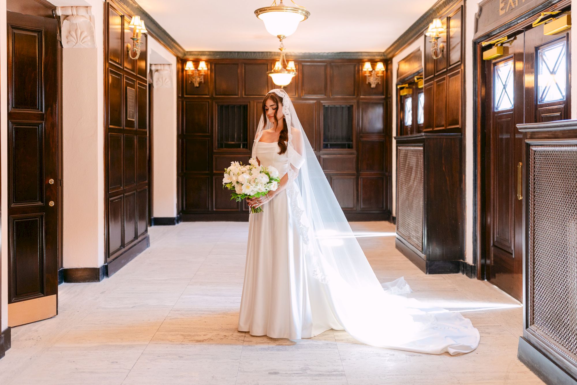 A bride in a white wedding gown with a long veil stands in a grand hallway, holding a bouquet of flowers. End.