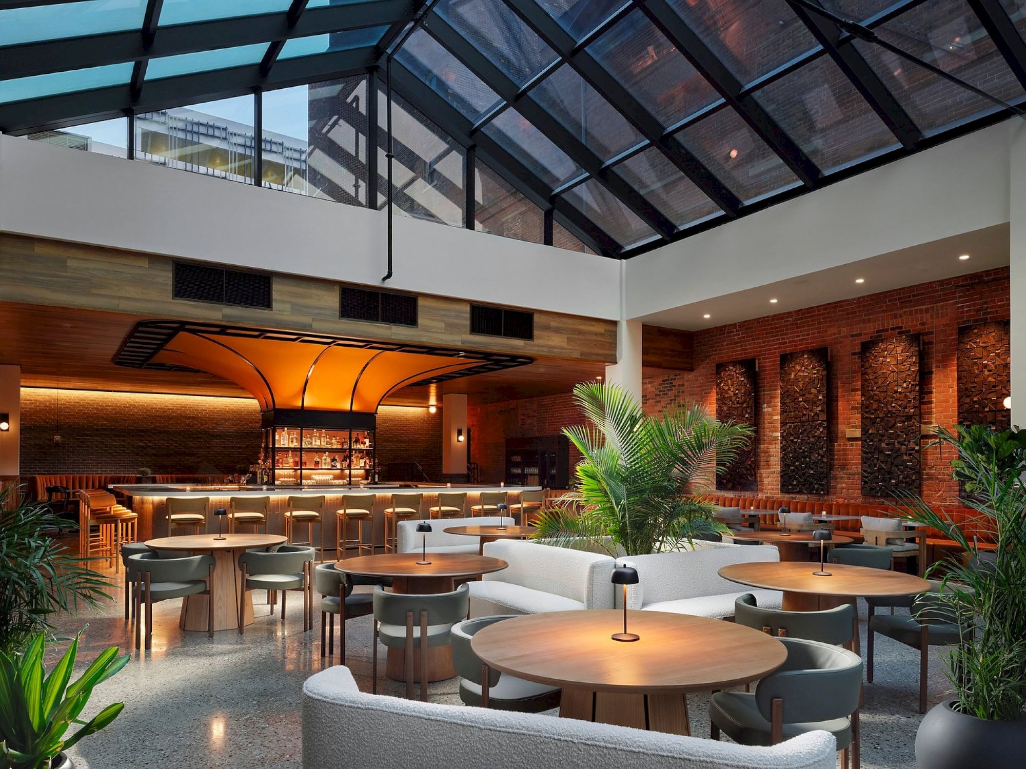 The image shows a modern lounge with a glass ceiling, wooden tables, and a bar. Green plants enhance the cozy and stylish ambiance.