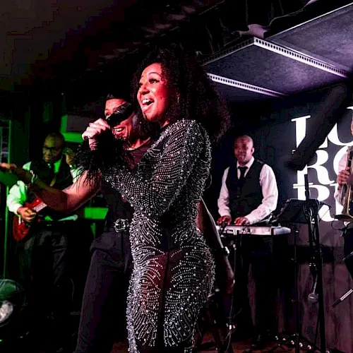 A singer in a sparkly jumpsuit performs live with a jazz band on stage under green lights.