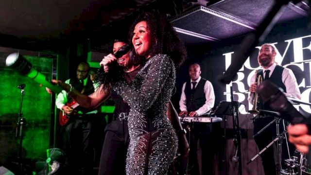A singer in a sparkly jumpsuit performs live with a jazz band on stage under green lights.
