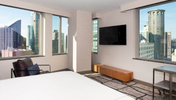 A modern hotel room with large windows showing a city skyline, a bed, TV, armchair, and a bench.