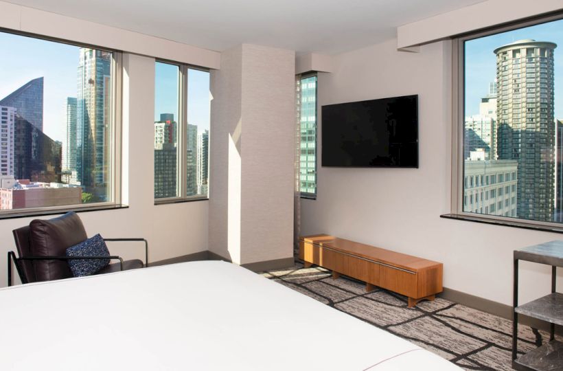 A modern hotel room with large windows showing a city skyline, a bed, TV, armchair, and a bench.