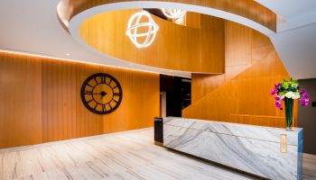 A modern lobby with a marble reception desk, wooden accents, spiral ceiling design, large wall clock, and contemporary lighting fixtures.