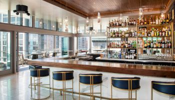 A modern bar with high stools, a well-stocked liquor shelf, and large windows offering a city view; elegant and inviting space.