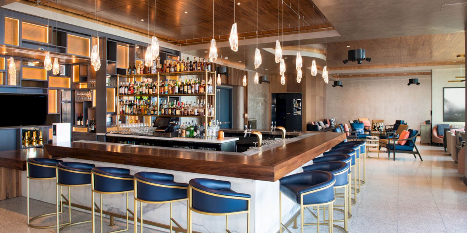 The image shows a modern bar with a large counter, stylish bar stools, hanging lights, and shelves stocked with bottles in a spacious setting.