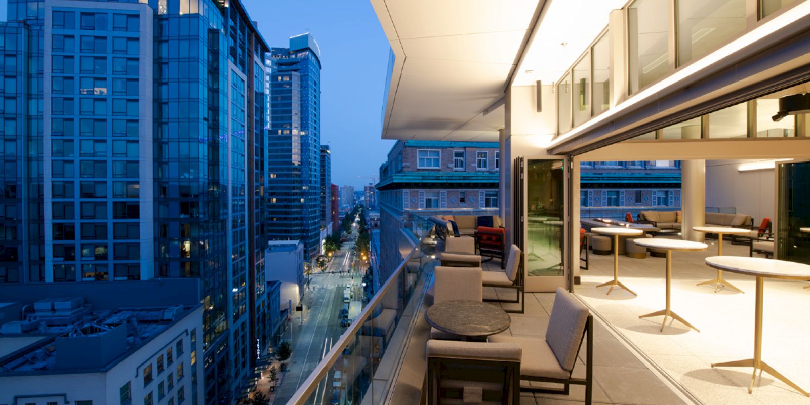 A modern rooftop terrace with seating overlooks a cityscape featuring tall buildings during the evening, with warm lighting enhancing the ambiance.