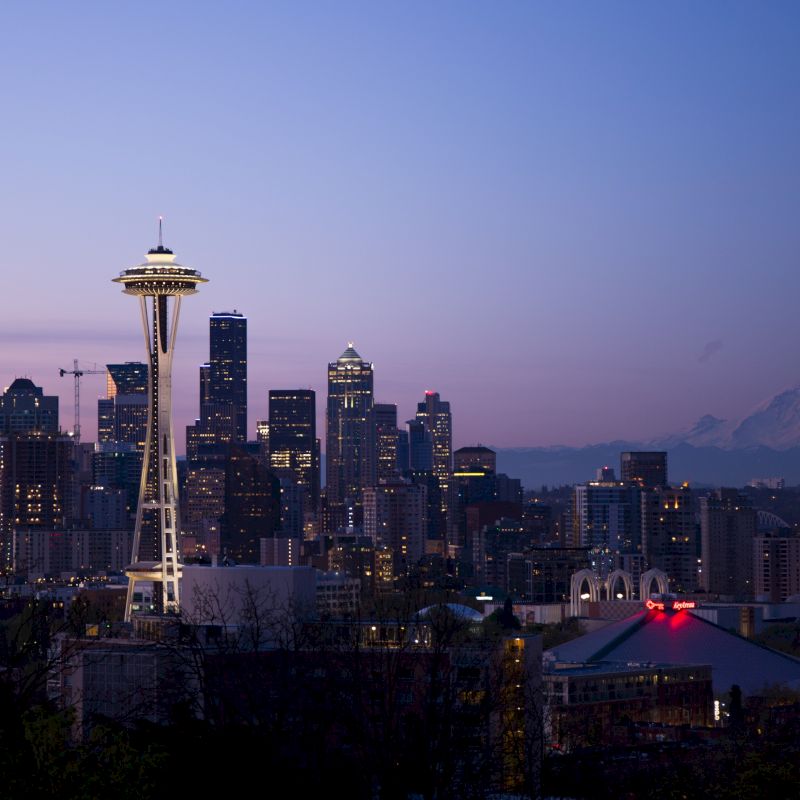 A cityscape of Seattle at dusk featuring the Space Needle, with mountain in the background and a clear sky.