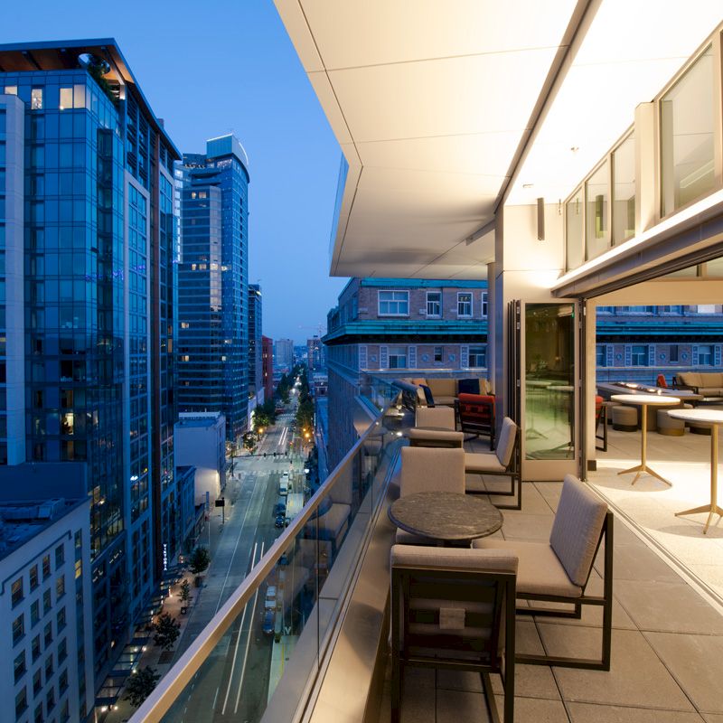 A modern outdoor terrace with seating overlooks a city street. Illuminated buildings are visible in the evening light.