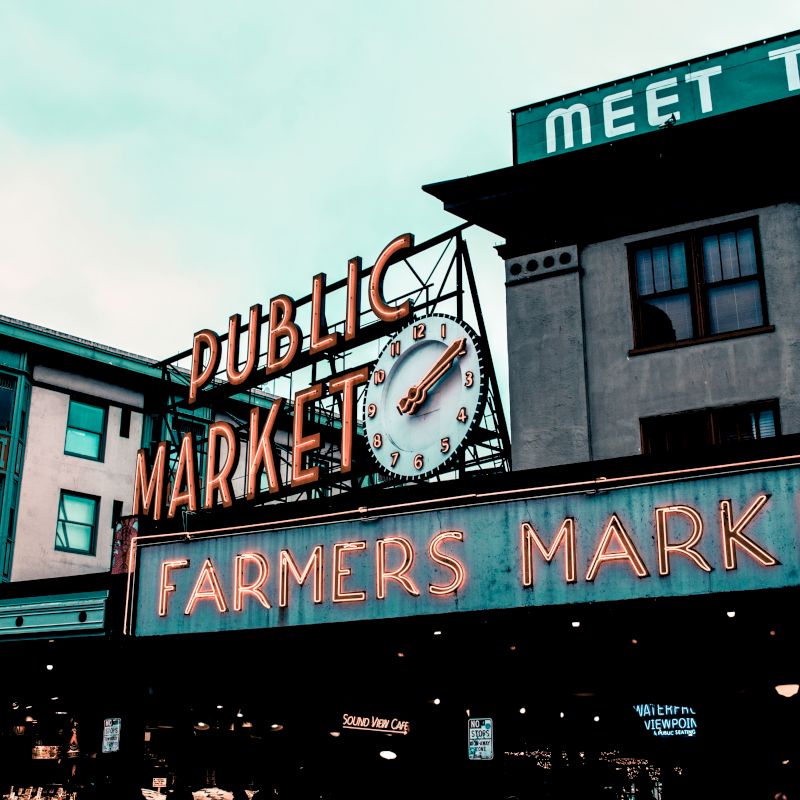 The image shows a classic neon sign for a public farmers market with a clock, in an urban setting with visible surrounding buildings.