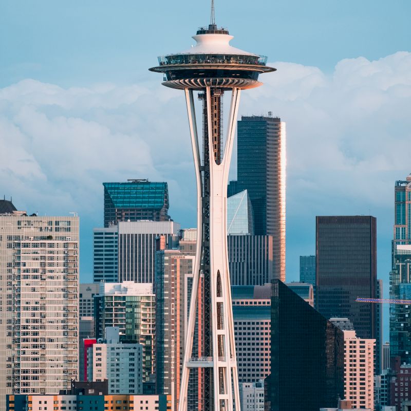 The image features the Seattle Space Needle with a backdrop of modern skyscrapers and cloud-filled sky, capturing the city's iconic skyline.