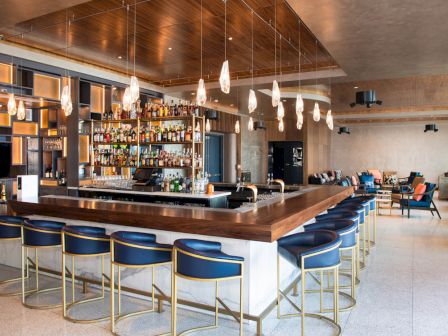 The image shows a modern bar with a central counter, stylish bar stools, and hanging lights in an upscale, cozy setting.