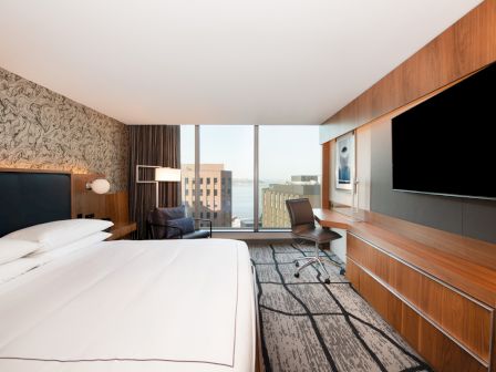 A modern hotel room with a large bed, TV, desk, chair, and a window with a city view, featuring sleek decor and a patterned carpet.