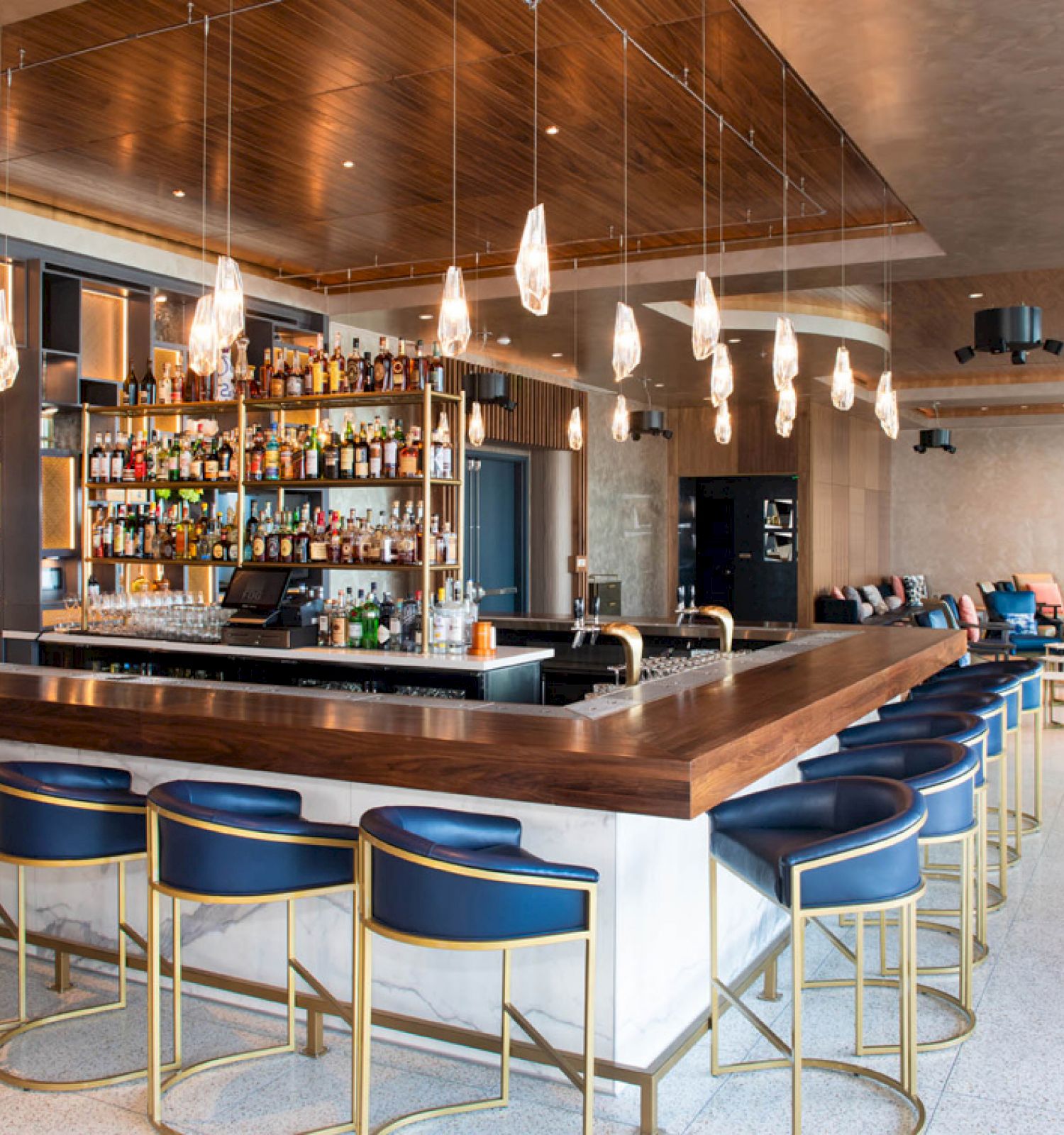 The image shows a stylish bar with a central island, blue stools, pendant lighting, and a variety of bottles displayed on shelves.