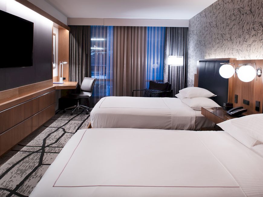 The image shows a hotel room with two beds, a TV, a desk, chairs, and lamps by the window, with patterned walls and carpet.