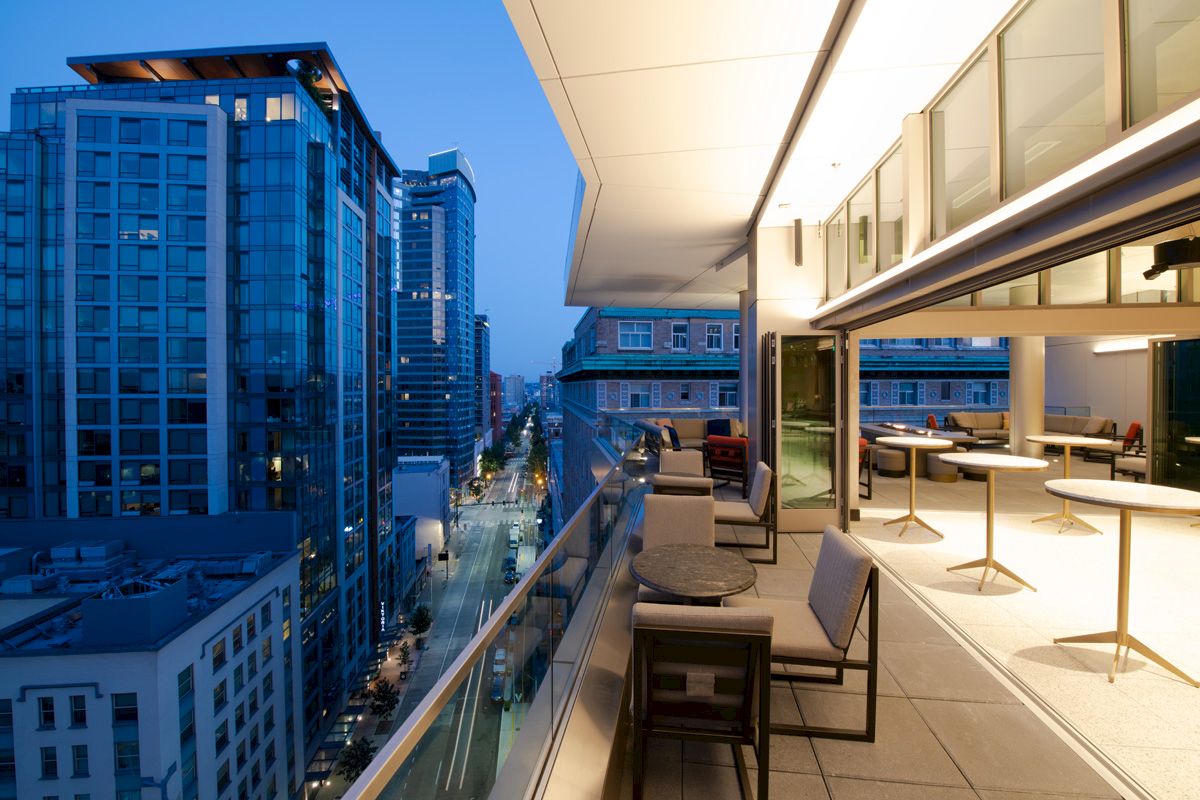 A modern rooftop terrace with seating overlooks a cityscape of tall buildings during evening, with a street view.