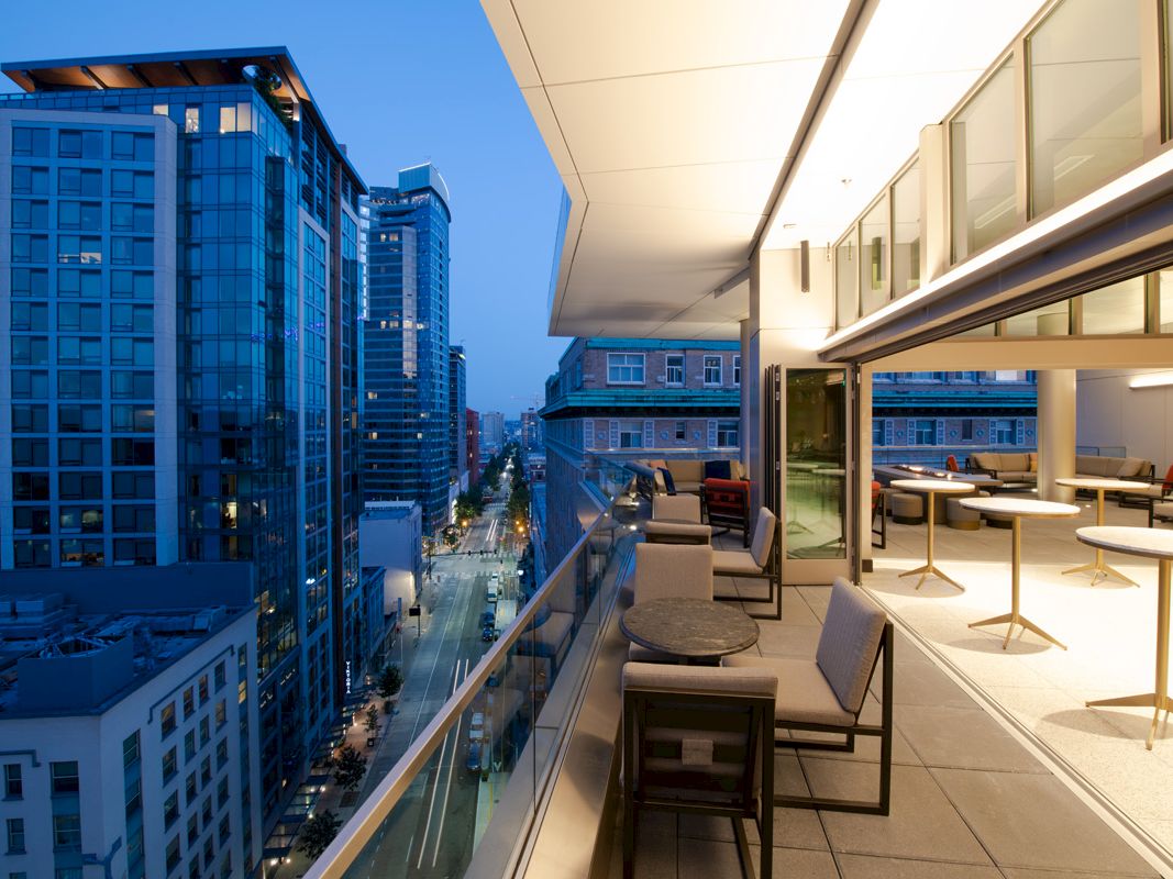 A modern rooftop terrace with seating overlooks a cityscape of tall buildings during evening, with a street view.