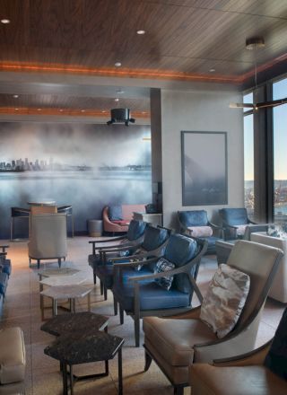 The image shows a modern lounge with stylish seating, large windows, and a scenic view of a body of water and cityscape.