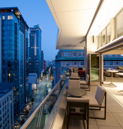 A modern rooftop terrace with seating overlooks a city skyline at dusk, featuring tall buildings and a street view.