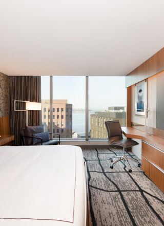 A modern hotel room with a large bed, flat-screen TV, desk, and chair. A city view is visible through the large window.