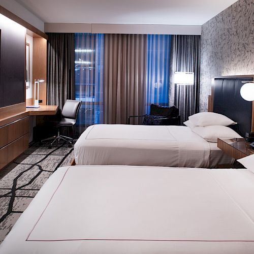 The image shows a modern hotel room with two beds, a flat-screen TV, a desk, and stylish lighting, featuring contemporary decor.