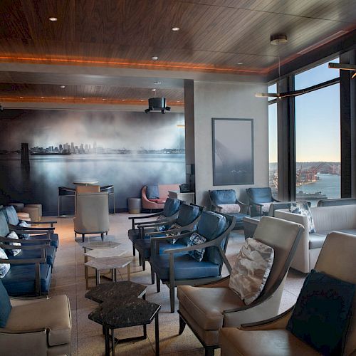 The image shows a modern lounge with leather chairs, tables, large windows offering a scenic view, and ambient lighting.