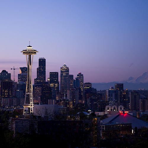 The image shows Seattle's skyline at dusk, featuring the Space Needle and Mount Rainier in the background against a dimly lit sky.