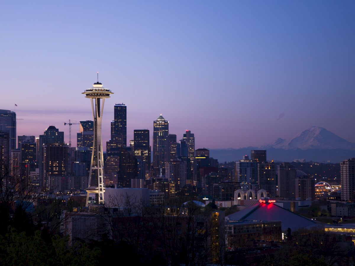 A city skyline at dusk featuring a prominent tower, with a mountain faintly visible in the background and buildings illuminated.
