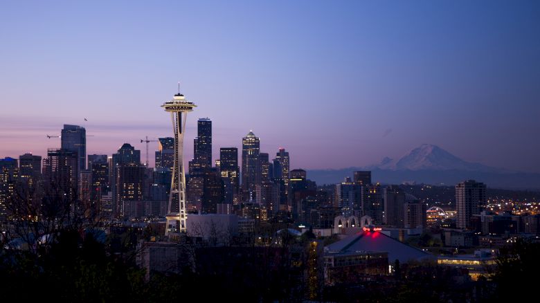 A city skyline at dusk featuring a prominent tower, with a mountain faintly visible in the background and buildings illuminated.