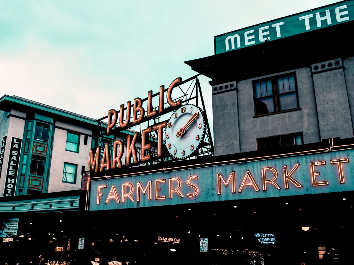 A neon sign for a public farmers market, featuring a clock, with surrounding buildings in an urban setting under a cloudy sky.
