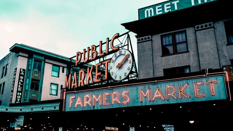 A neon sign for a public farmers market, featuring a clock, with surrounding buildings in an urban setting under a cloudy sky.