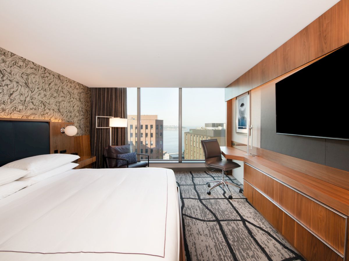 A modern hotel room with a large bed, wall-mounted TV, desk, and a window view of buildings, featuring stylish decor and lighting.