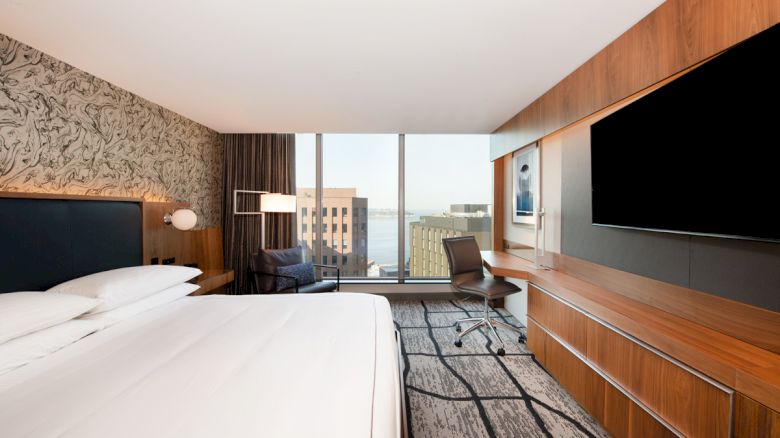 A modern hotel room with a large bed, wall-mounted TV, desk, and a window view of buildings, featuring stylish decor and lighting.