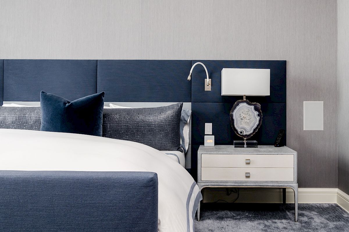 A modern bedroom with a blue and white theme, featuring a bed, a nightstand with a lamp, and a decorative pillow.