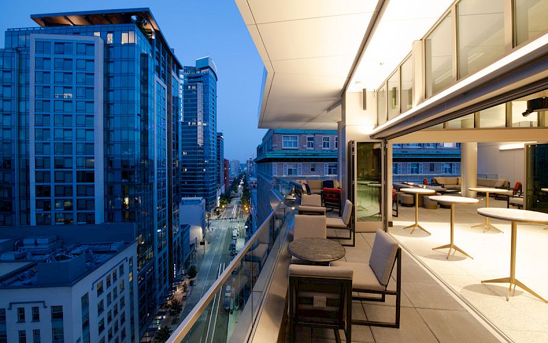 A modern rooftop terrace with seating overlooks a cityscape of tall buildings as dusk falls, creating a warm and inviting atmosphere.