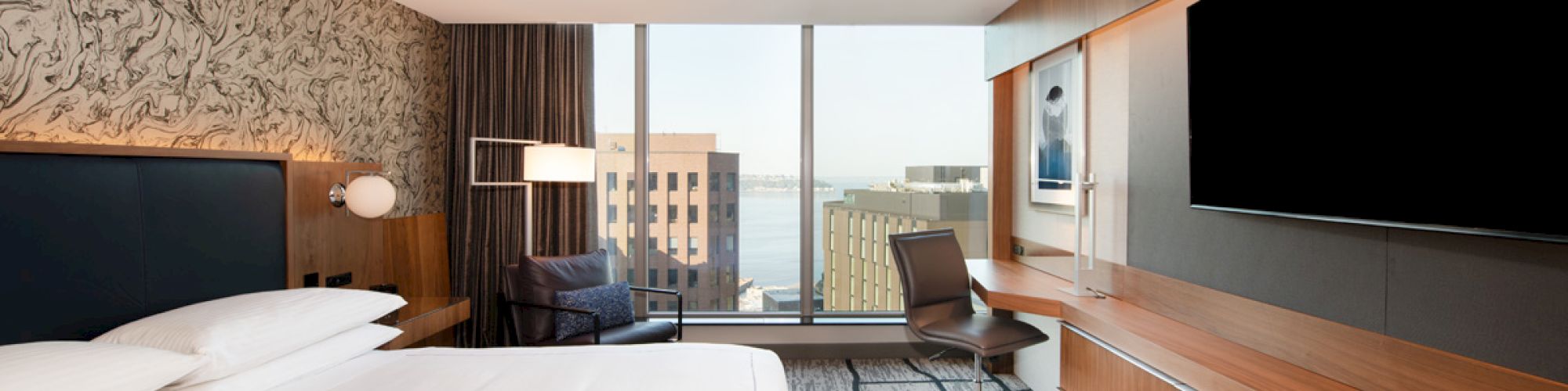A modern hotel room with a large bed, wall-mounted TV, city view window, desk, and stylish decor.