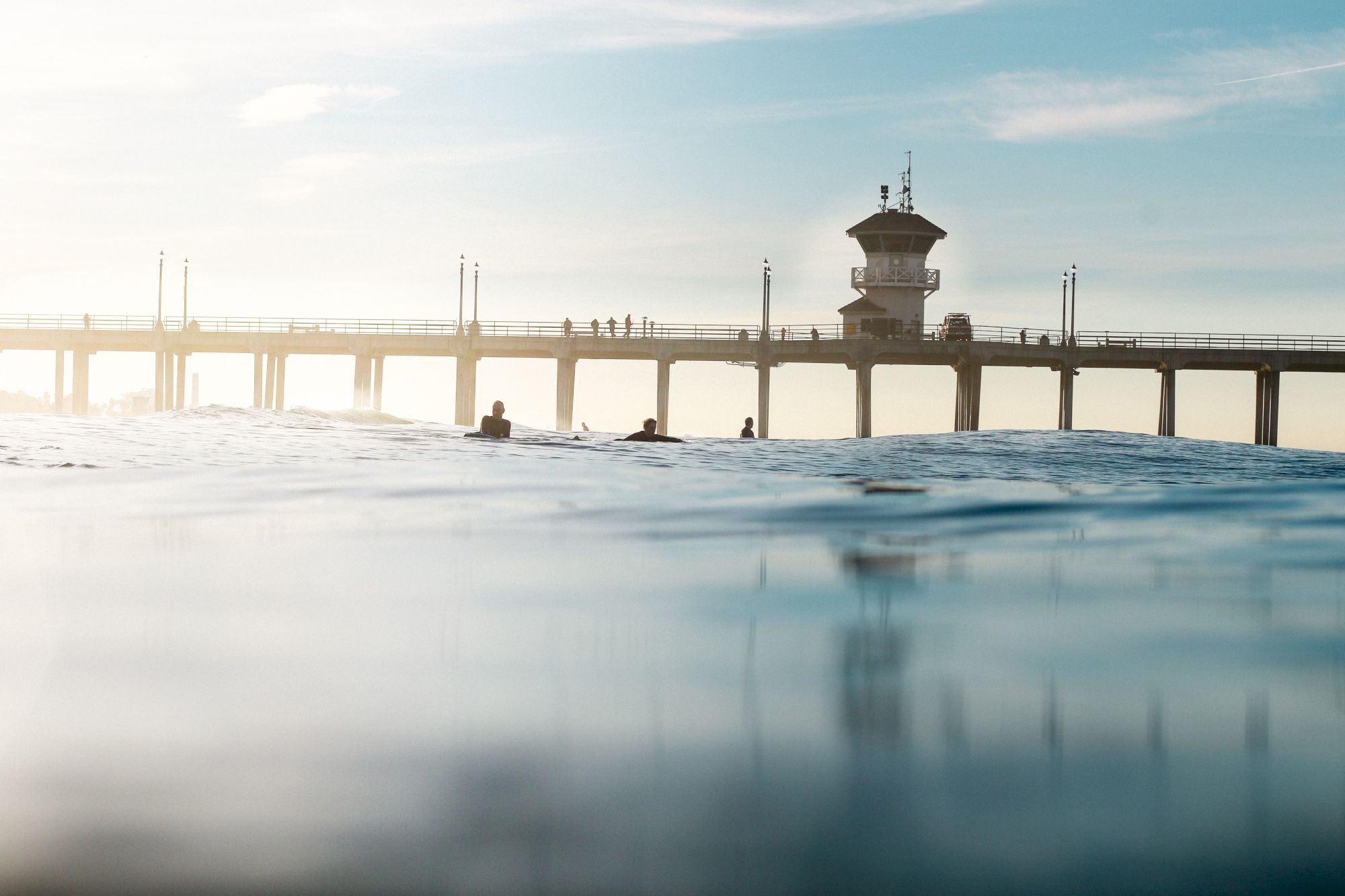 A pier stretches across calm water with people walking on it, under a clear sky. Silhouetted surfers float near the foreground in the water.